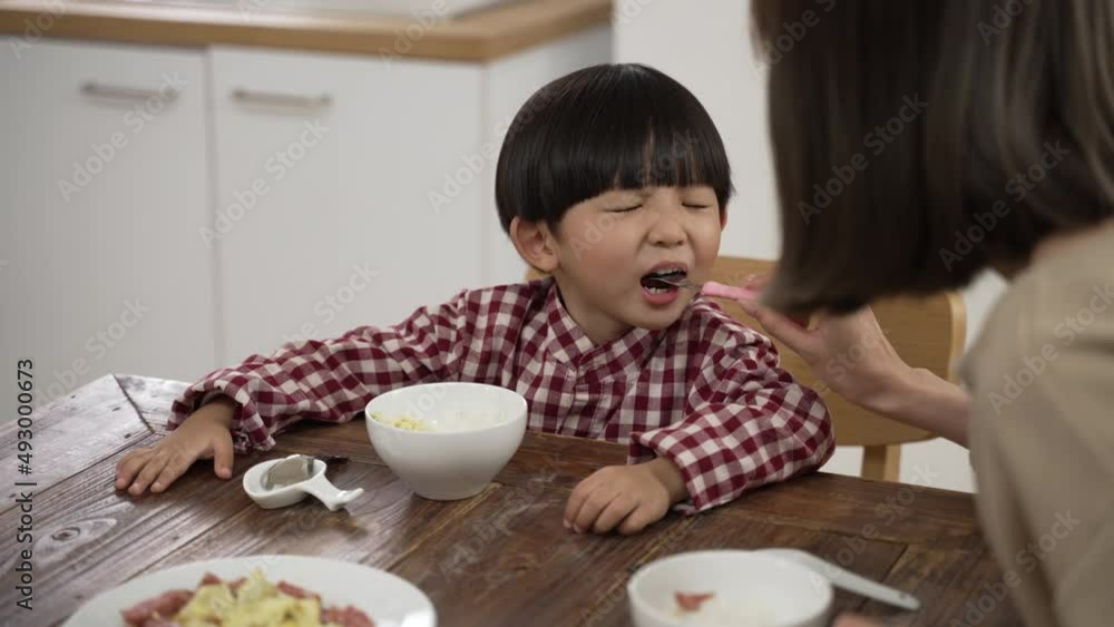 slow motion with selective focus of cute Asian little boy opening his mouth as his mother feeds him a piece of meat with a fork at dining table. slow motion with selective focus of cute Asian little boy opening his mouth as his mother feeds him a piece of meat with a fork at dining table.