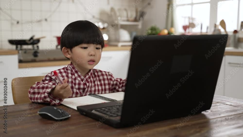 Asian home learning boy practicing speaking with teacher while taking virtual class with computer and writing notes at dining table Asian home learning boy practicing speaking with teacher while taking virtual class with computer and writing notes at dining table