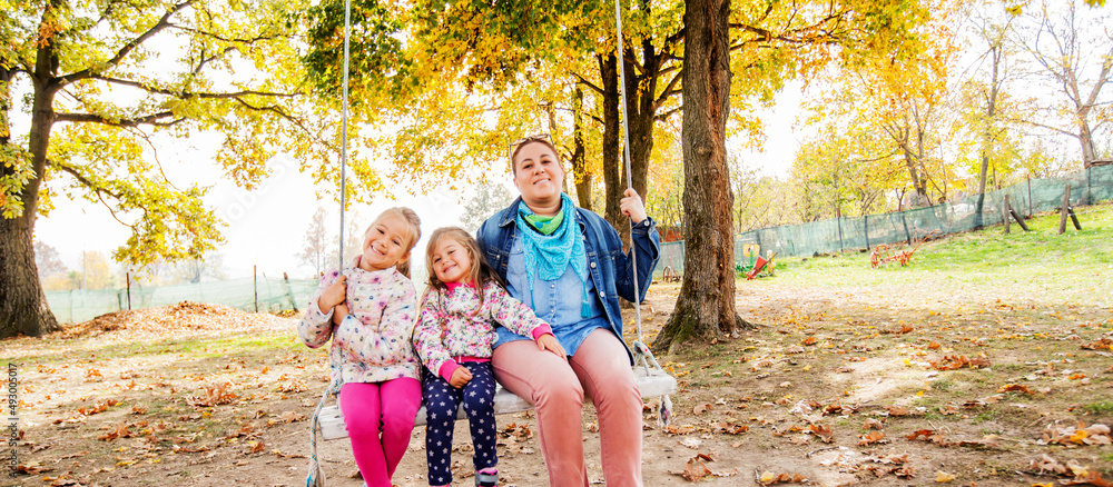 Fototapeta premium Happy Family Relaxing On Swing In Park. Smiling mother with daughters fun on sunny autumn day in nature.