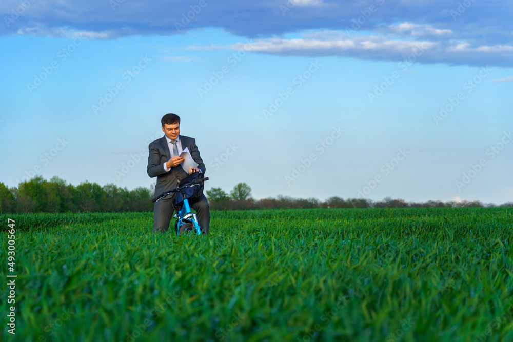 businessman dressed in a business suit, poses with bicycle in green grass field and reads documents, beautiful nature in spring, business concept