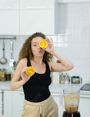 vegetarian woman happy funny moment by hiding one eye behind half orange while cooking fruit juice in kitchen. beautiful caucasian woman cooking healthy diet fruit or vegetable in kitchen