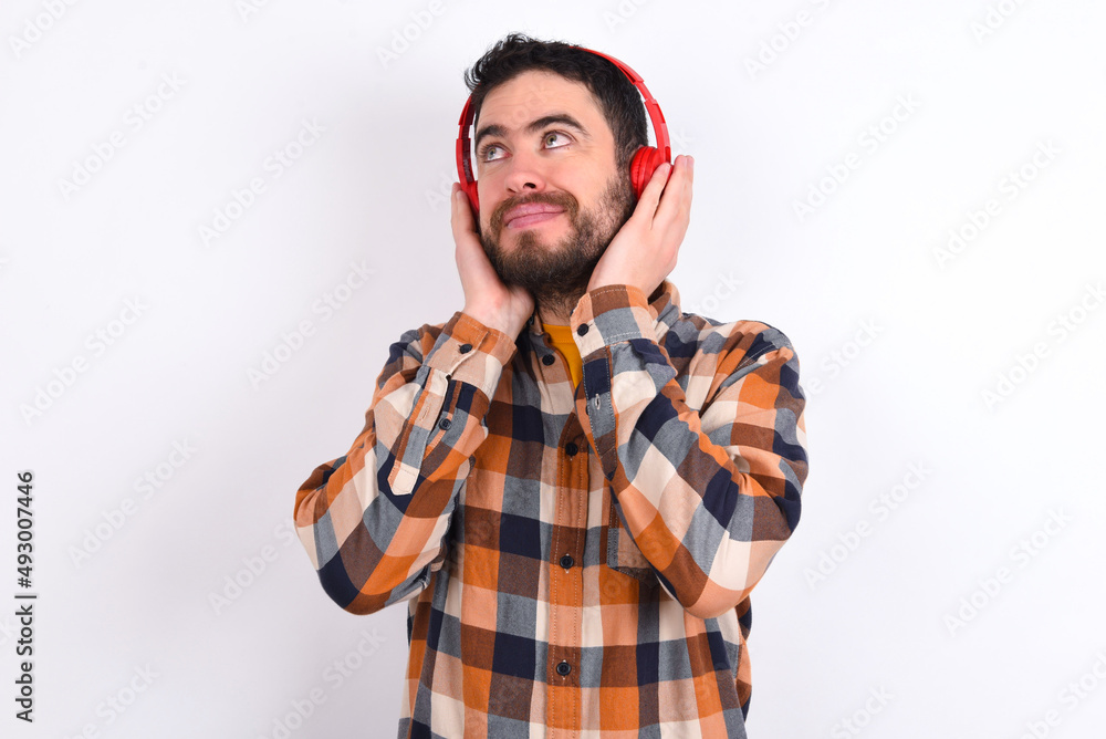 young caucasian man wearing plaid shirt over white background wears stereo headphones listens music concentrated aside. People hobby lifestyle concept young caucasian man wearing plaid shirt over white background wears stereo headphones listens music concentrated aside. People hobby lifestyle concept