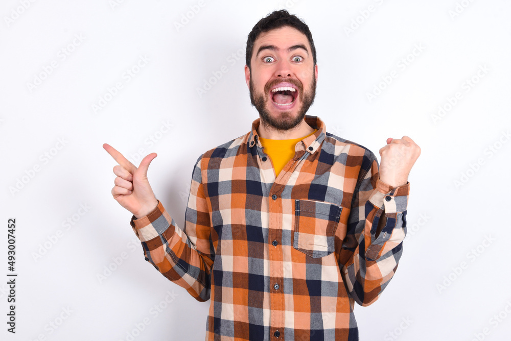 young caucasian man wearing plaid shirt over white background points at empty space holding fist up, winner gesture. young caucasian man wearing plaid shirt over white background points at empty space holding fist up, winner gesture.