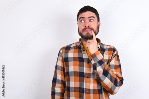 Lovely dreamy young caucasian man wearing plaid shirt over white background keeps finger near lips looks aside copy space.