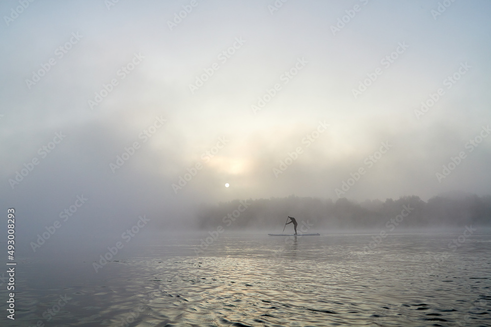 Silhouette of man paddles on standing up paddle board (SUP) on misty foggy river. Enjoying nature and solitude in weightlessness