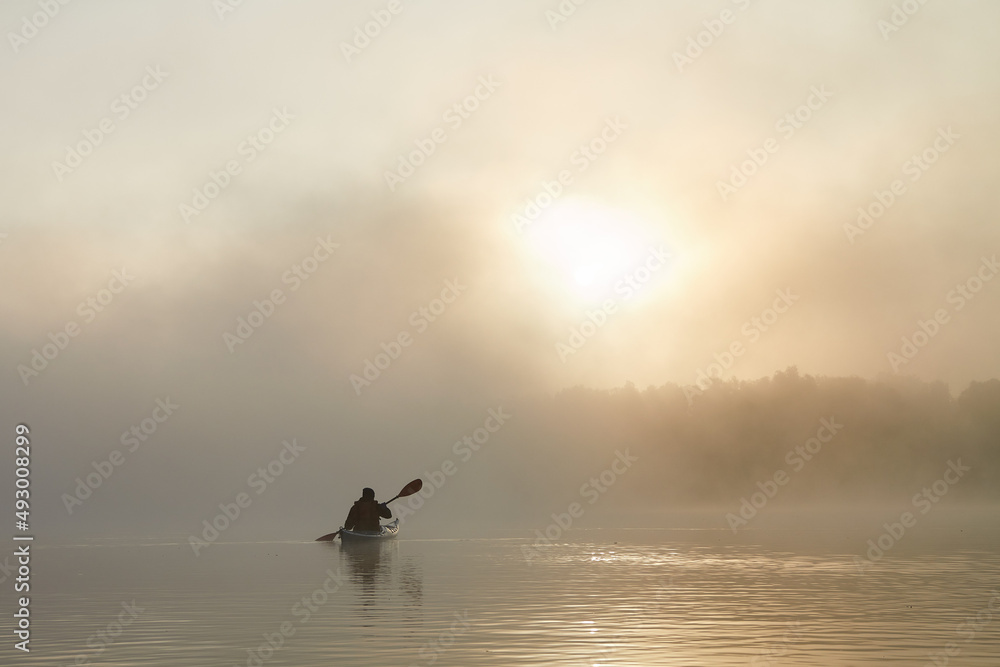 Fototapeta premium Man in kayak paddling through dense fog in wilderness area at autumn sunrise