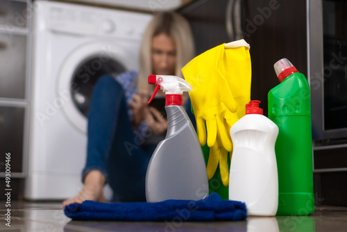 Close-up of bottles of cleaning products, rags, yellow rubber gloves, in the background a young woman with a mobile phone in her hands during a break in cleaning the house