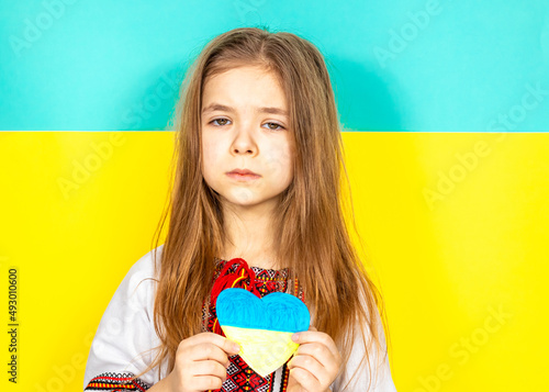 A girl in a national Ukrainian vyshyvanka holds a heart painted in yellow and blue tones as a sign of love for Ukraine, close-up against the background of the Ukrainian flag. The concept of peace in photo