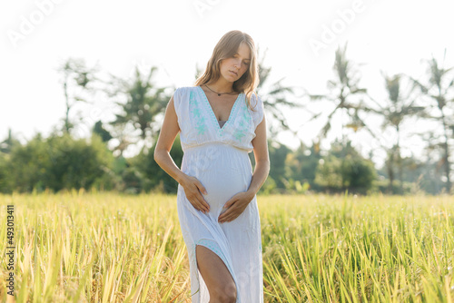 Pregnant woman stand at the field with green grass and embrace her belly. Sunshine summer vibes