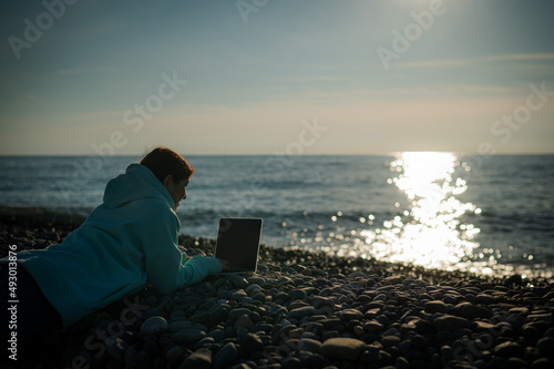 Caucasian woman lying on her stomach on a pebble beach and typing on a laptop. Remote work freelancer. © Михаил Решетников