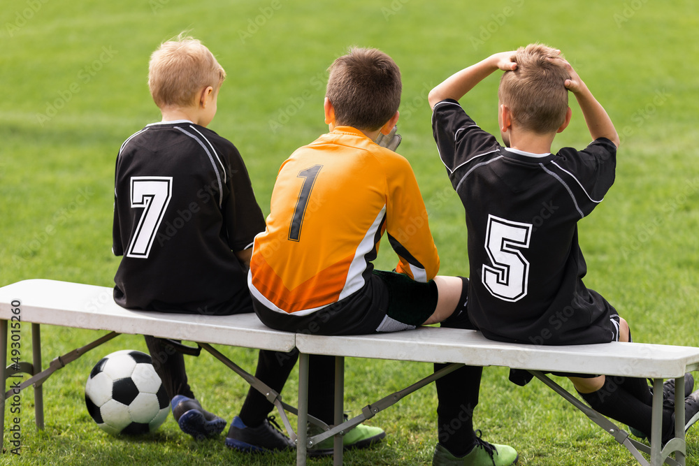 Obraz premium Soccer players sitting on sideline bench. School football team. Youth soccer players sitting together on substitute bench and watching match