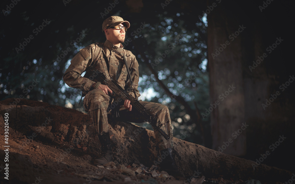 young soldier of ukraine with a backpack on his back in a gray camouflage uniform, in tactical glasses and in a cap with a machine gun in his hands sits on the ground in a destroyed hospital building