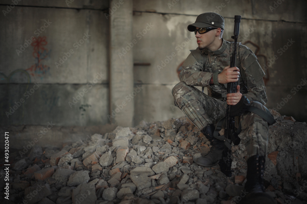 a military man with a machine gun in his hands in a gray camouflage uniform, wearing a cap and tactical glasses sits on the ground inside a large destroyed building a military man with a machine gun in his hands in a gray camouflage uniform, wearing a cap and tactical glasses sits on the ground inside a large destroyed building