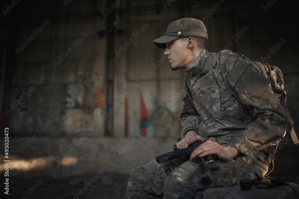 a soldier with a machine gun in a gray camouflage uniform wearing a cap and tactical black glasses sits on a concrete floor inside a large destroyed building a soldier with a machine gun in a gray camouflage uniform wearing a cap and tactical black glasses sits on a concrete floor inside a large destroyed building