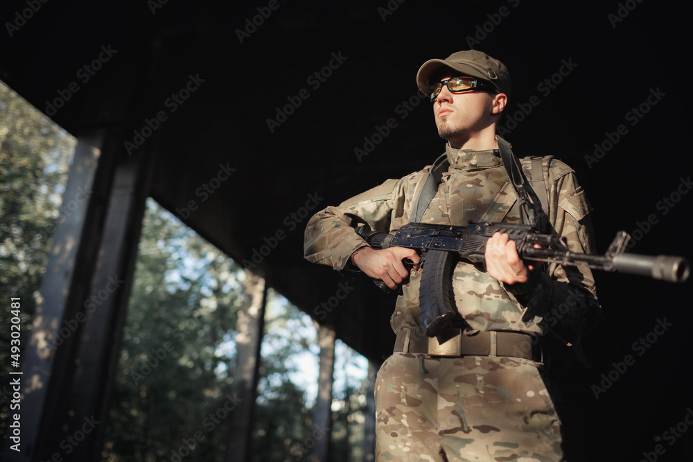 a serious male soldier in a gray camouflage uniform, wearing tactical glasses with yellow lenses and a cap with a black machine gun in his hands inside a large authorized factory