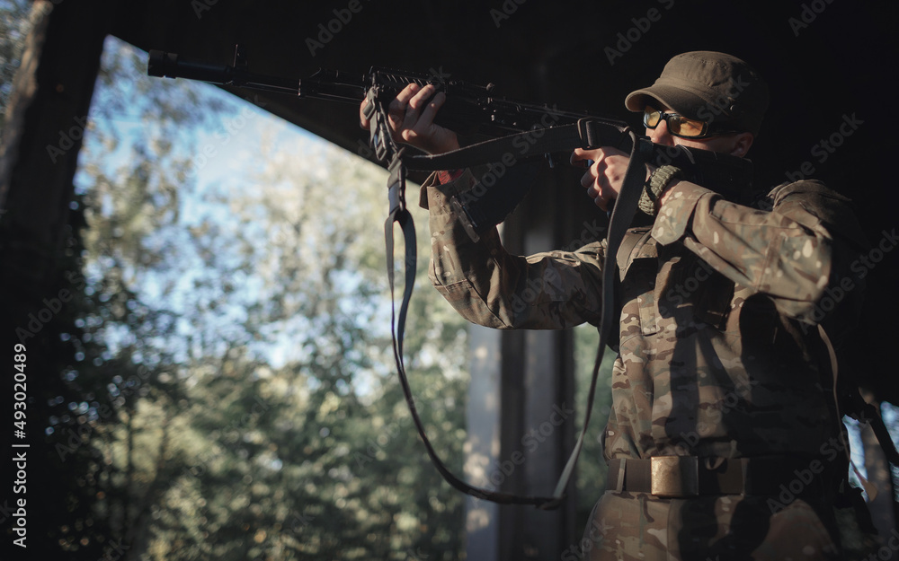 Naklejka premium a young soldier defender of his country in a camouflage gray uniform, wearing tactical black glasses and a cap with a machine gun in his hands aiming into the distance inside a large building