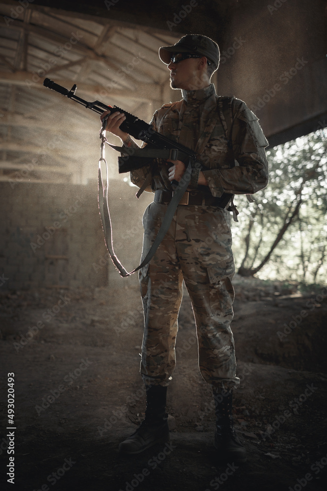a silhouette of a soldier in a camouflage protective uniform, in tactical black glasses and in a cap with a machine gun in his hands stands inside a large permitted house a silhouette of a soldier in a camouflage protective uniform, in tactical black glasses and in a cap with a machine gun in his hands stands inside a large permitted house