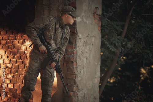a soldier with combat experience in a gray camouflage uniform, wearing tactical glasses and a cap with a machine gun in his hands, peeks around the corner in search of the enemy