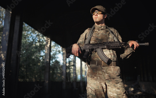 young stern male soldier in a gray camouflage uniform, wearing tactical goggles with yellow lenses and a cap with a black machine gun in his hands inside a large authorized factory