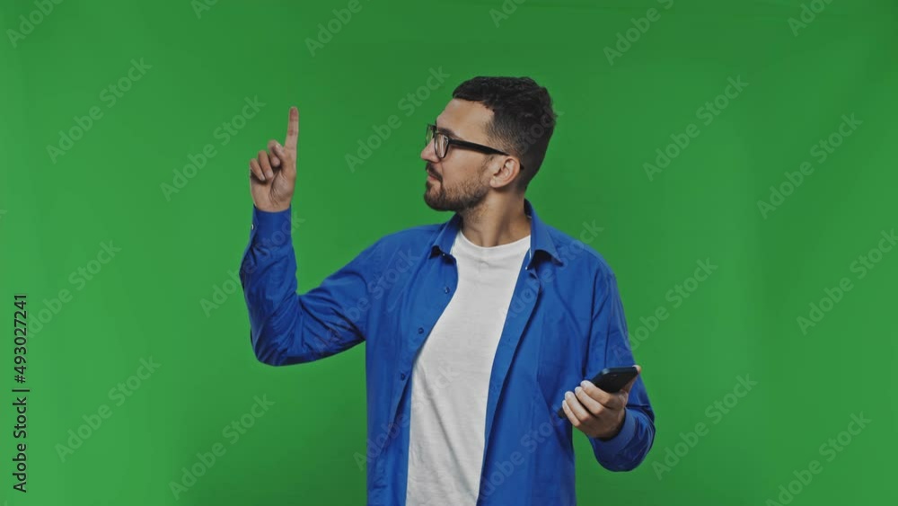 Bearded man wesring eyeglasses uses a smartphone and points up on copy space with smilling face on isolated green background Bearded man wesring eyeglasses uses a smartphone and points up on copy space with smilling face on isolated green background