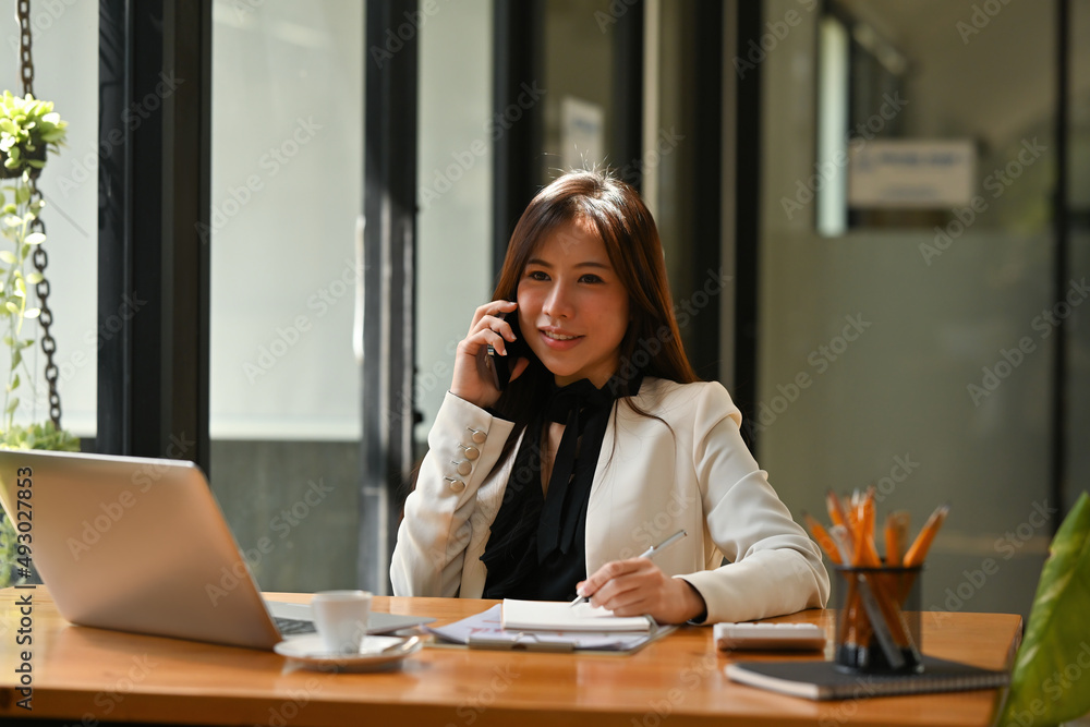 A portrait of a young pretty Asian woman sitting in the office using a telephone, laptop, calculator, working on the documents, for business, finance and technology concept. A portrait of a young pretty Asian woman sitting in the office using a telephone, laptop, calculator, working on the documents, for business, finance and technology concept.
