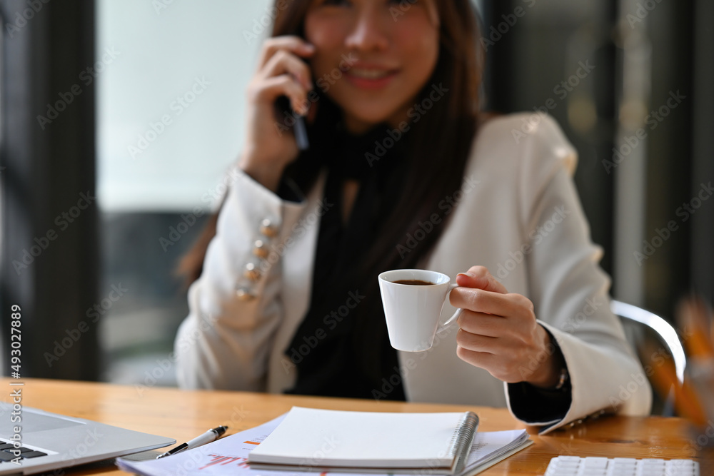 Cropped view young pretty Asian woman sitting in the office using a telephone and holding a cup of coffee, working on the documents, for business, finance and technology concept. Cropped view young pretty Asian woman sitting in the office using a telephone and holding a cup of coffee, working on the documents, for business, finance and technology concept.