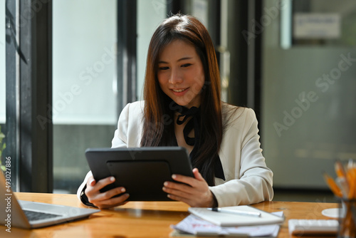 A portrait of a young pretty Asian woman sitting in the office using a tablet, calculator, working on data, charts and documents on the table, for business, finance and technology concept.