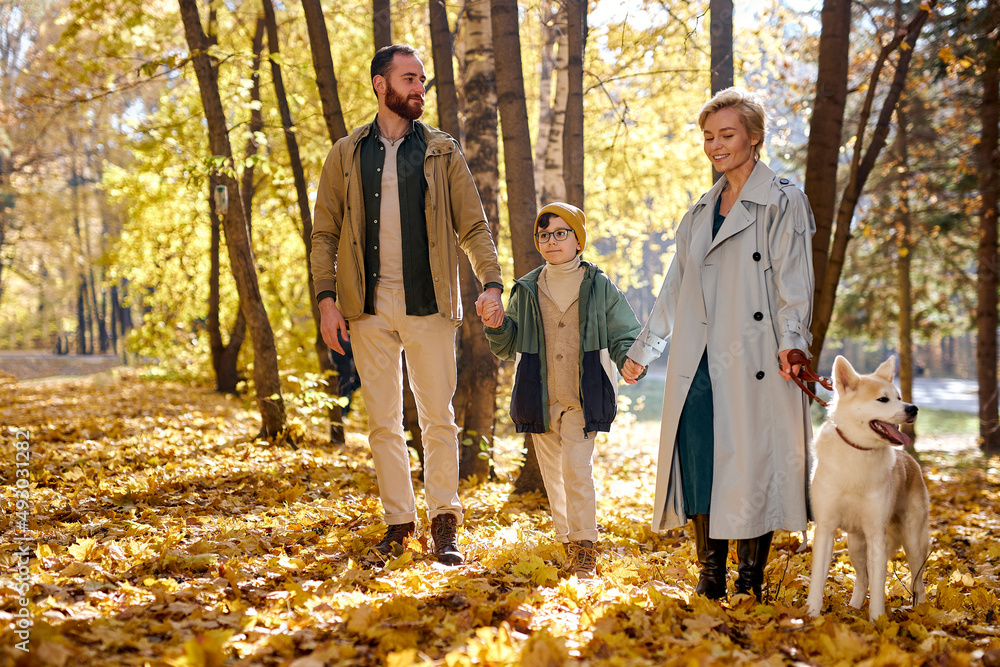 Fototapeta premium caucasian family with child and dog on walk in autumn forest, lead dog on a leash. Family walk with white dog in park. attractive man, woman and kid boy spend time together, love their pet