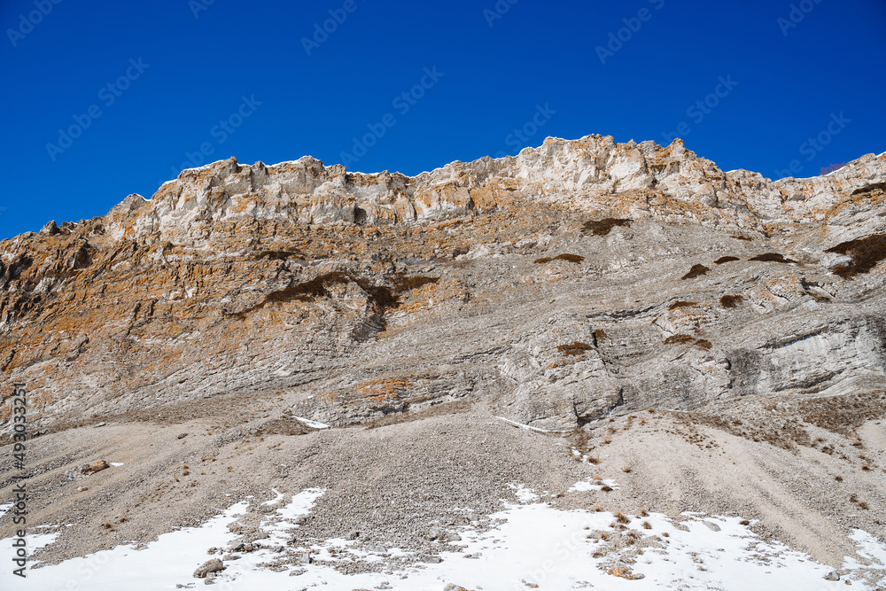 The high cliffs are grey. The top of a stone mountain against the sky, snow fell at the foot of the mountains. View from below of the high mountains.