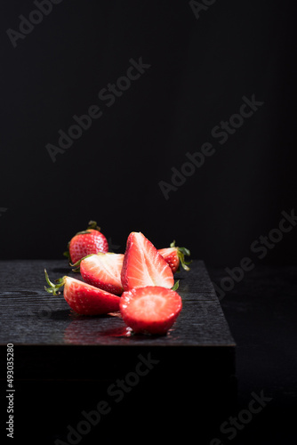 Fresas rojas enteras y cortadas en una mesa negra  sobre un fondo negro.