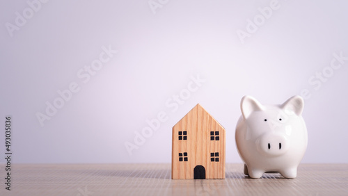 White piggy bank with wooden house on wood board table with gray background. Concept for financial home loan or money saving for house buying.