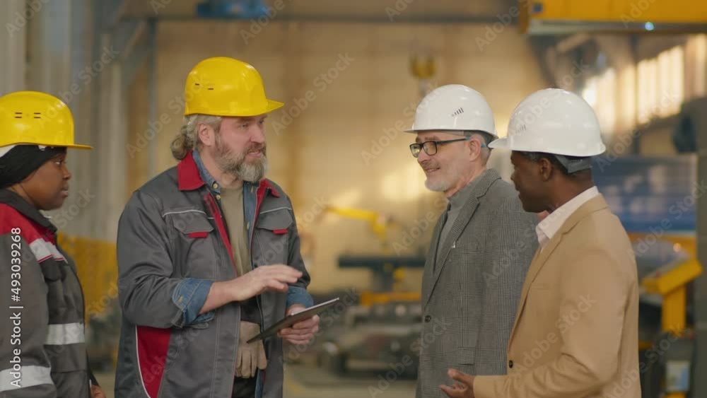Mature engineer and technician in hardhats shaking hands and discussing something on digital tablet with multiethnic colleagues while standing together in factory Mature engineer and technician in hardhats shaking hands and discussing something on digital tablet with multiethnic colleagues while standing together in factory