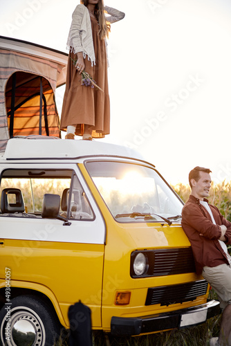 Beautiful caucasian couple spend time in nature, came there on mini van, woman is standing on roof of yellow van while man is standing next to van. hippie life, travel, holidays at sumemr photo