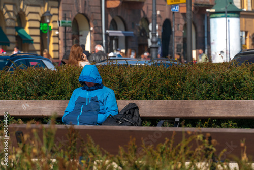 A lonely homeless man with a backpack sleeps sitting on a bench in a city park, refugees on the streets of European cities