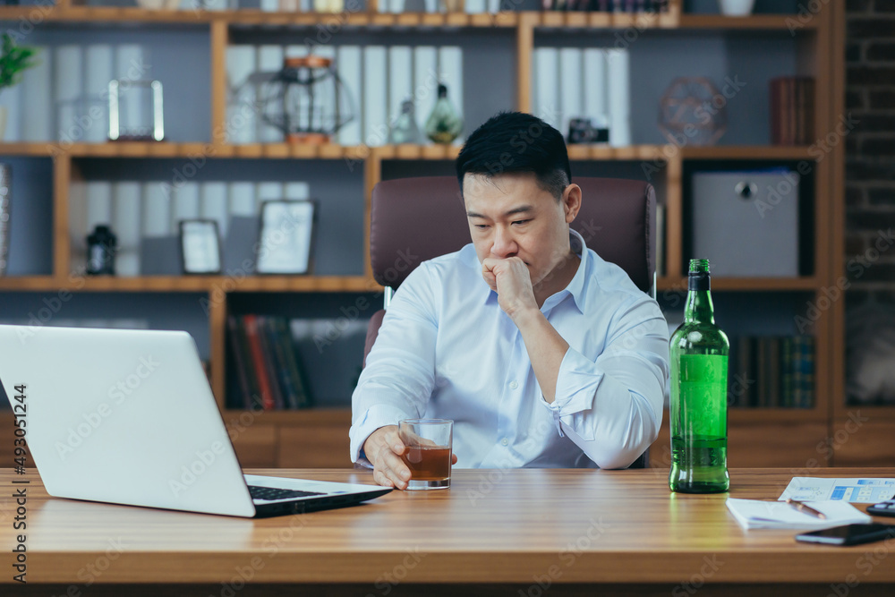 Asian businessman fails, man in despair drinks hard liquor, sitting late at the table in the office, depressed and hopeless