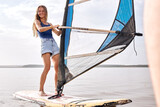 athlete caucasian fit female in shorts stands on windsurf and holds the sail. Windsurfing on lake water, relaxing on water.Summer water sports activities, recreation and travel concept