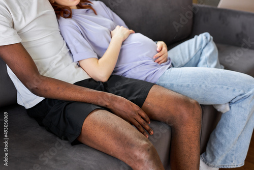 Cropped Pregnant woman, black man lying sitting on sofa having rest, leisure. Attractive lovely young couple enjoy rest spare time at weekends holidays in cozy living room. Natural light