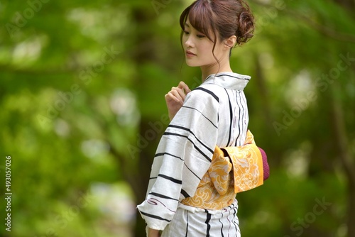 神社の境内にて緑の森林を背景にポーズをとる浴衣姿の若く美しい女性 