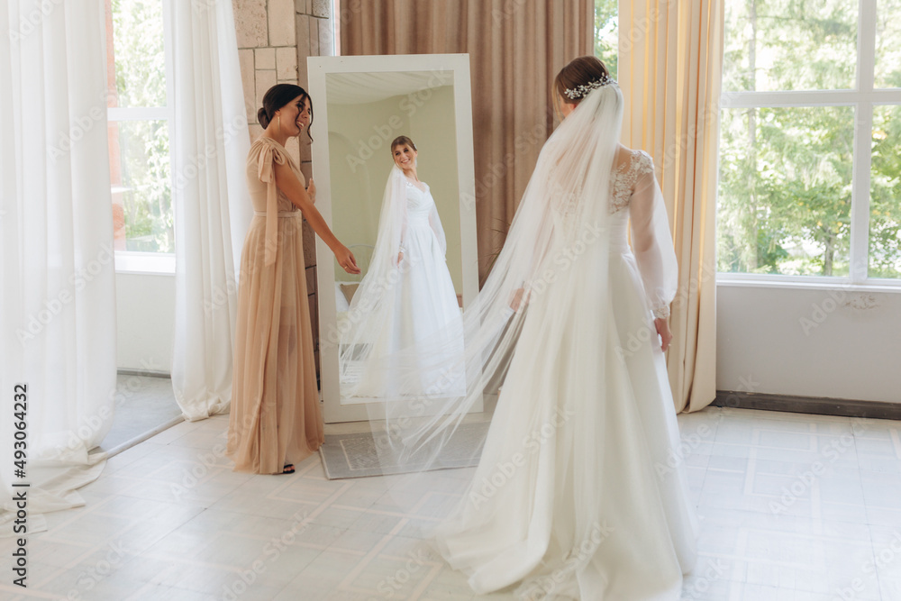 Side view of two happy smiling girls preparing for wedding in the morning. The young bride stands next to her friend, looking at her dress in the mirror in the bedroom. Side view of two happy smiling girls preparing for wedding in the morning. The young bride stands next to her friend, looking at her dress in the mirror in the bedroom.