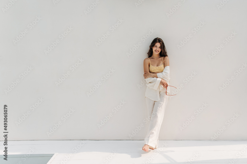 Full-length, sophisticated young european girl leaning against snow-white background with space for text. Brunette in casual summer clothes stands with her arms and legs crossed. Good morning concept Full-length, sophisticated young european girl leaning against snow-white background with space for text. Brunette in casual summer clothes stands with her arms and legs crossed. Good morning concept