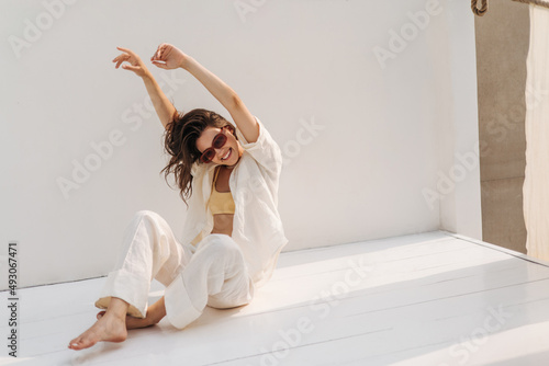 Cheerful young fair-skinned girl fooling around, waving arms and hair, sitting on floor near white background. Brunette in sunglasses is having fun alone. Concept of enjoying moments