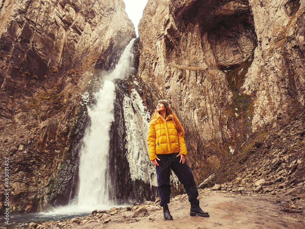 A woman looks up while standing against the background of an icy stormy waterfall flowing from a gorge of sheer rocks in the Caucasian highlands