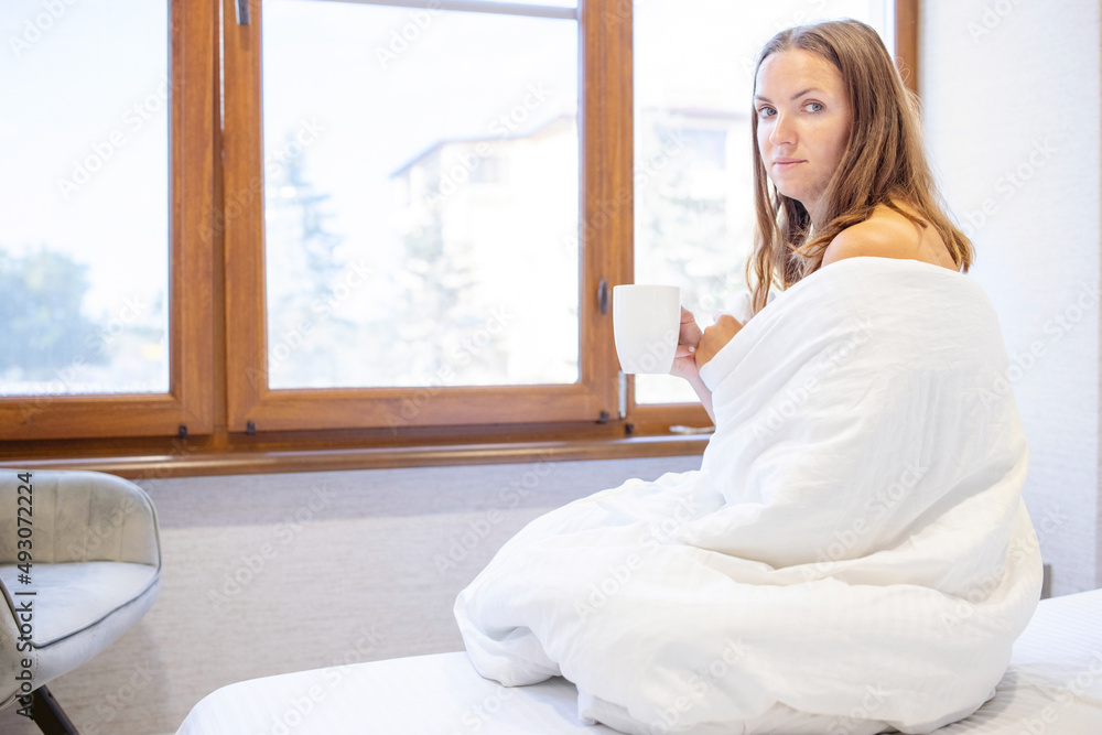 Close up of a woman sitting on bed holding a cup of coffee. Woman with bed coffee in hand sitting on bed. Morning coffee or tea in bed Close up of a woman sitting on bed holding a cup of coffee. Woman with bed coffee in hand sitting on bed. Morning coffee or tea in bed