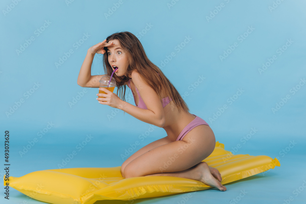Swimming is fun. Positive caucasian girl wearing in bikini in sunglasses going to poll with yellow inflatable mattress, blue studio background.