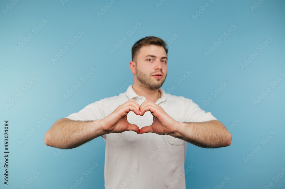 Adult man with bristles and in a white T-shirt stands on a blue background and shows a heart gesture to the camera. Adult man with bristles and in a white T-shirt stands on a blue background and shows a heart gesture to the camera.
