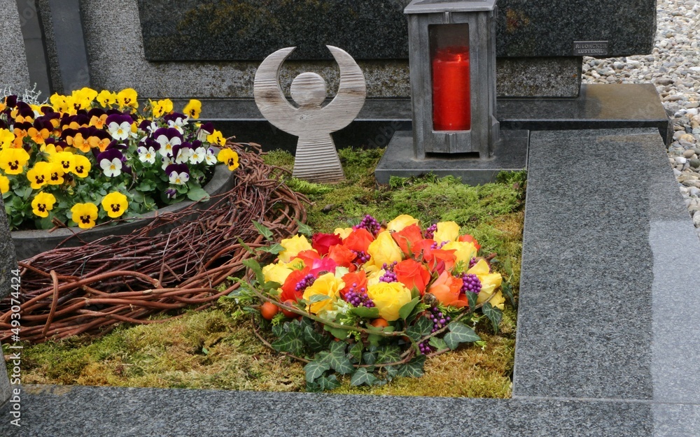 Mit Blumen geschmückte Gräber an einem katholischen Friedhof in Österreich, ein Blumengebinde mit Rosen, daneben eine Blumenschale mit Stiefmütterchen und ein Engel und eine Friedhofslaterne