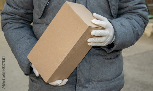 a man wearing white surgical gloves and grey winter jacket (winter coat) is holding a brown delivery box in hands, standing in front of a apartments door. outside. delivery service, products, goods,  © Alexandra