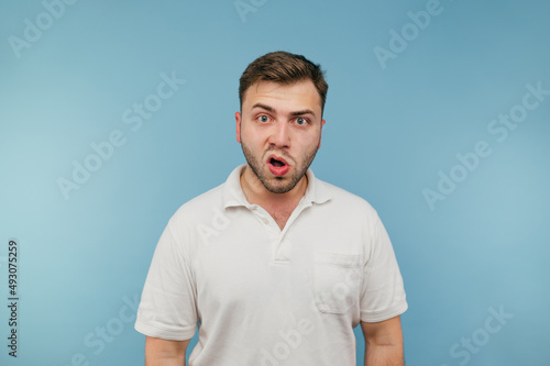 фотографія Surprised man with bristles on a blue background looks at the camera with a shocked face wearing a white T-shirt