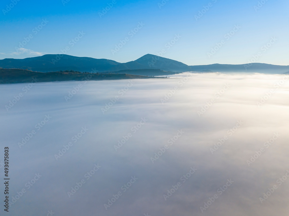 Flight over fog in Ukrainian Carpathians in summer. Mountains on the horizon. A thick layer of fog covers the mountains with a continuous carpet. Aerial drone view.