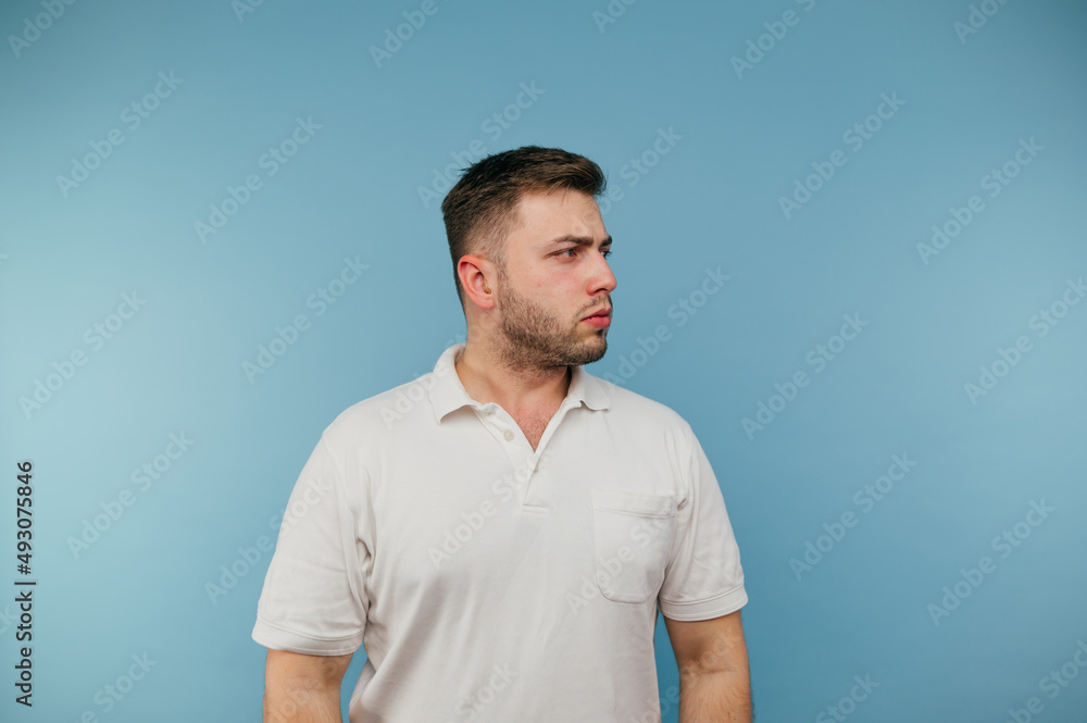 An adult man with bristles in a white T-shirt stands on a blue background and looks away with a serious face. An adult man with bristles in a white T-shirt stands on a blue background and looks away with a serious face.